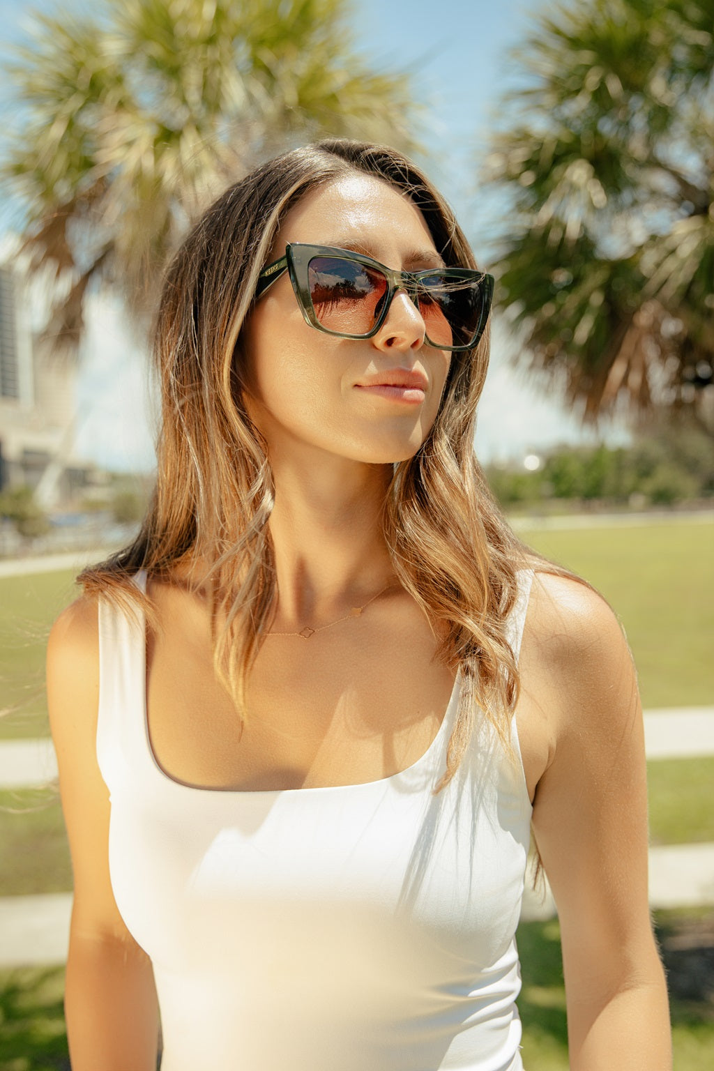 Woman wearing sunglasses and a white top in a park setting with palm trees.